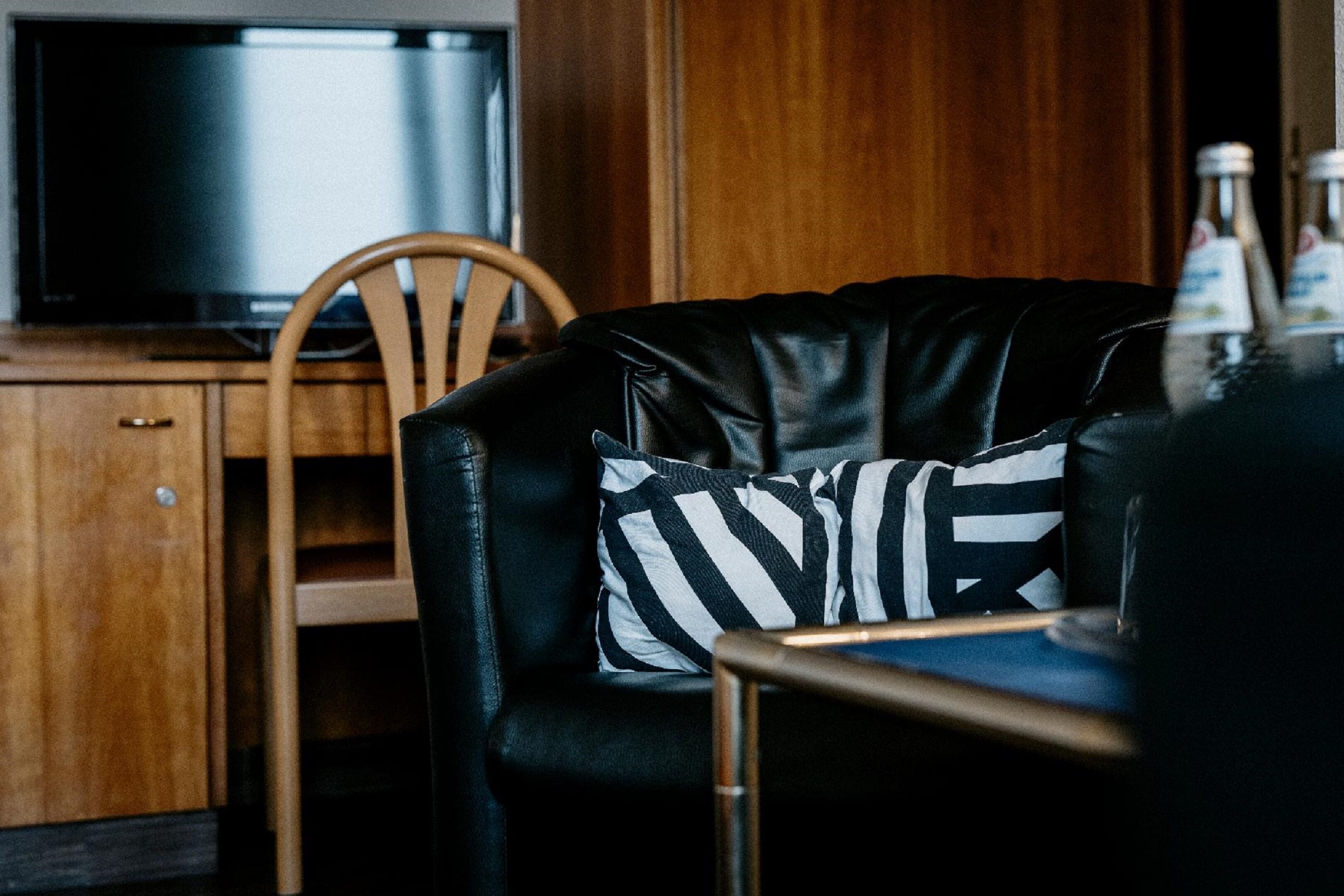 Een zwarte leren bank met een kussen met zwart-wit strepen, een houten stoel en een televisie op een houten kast.