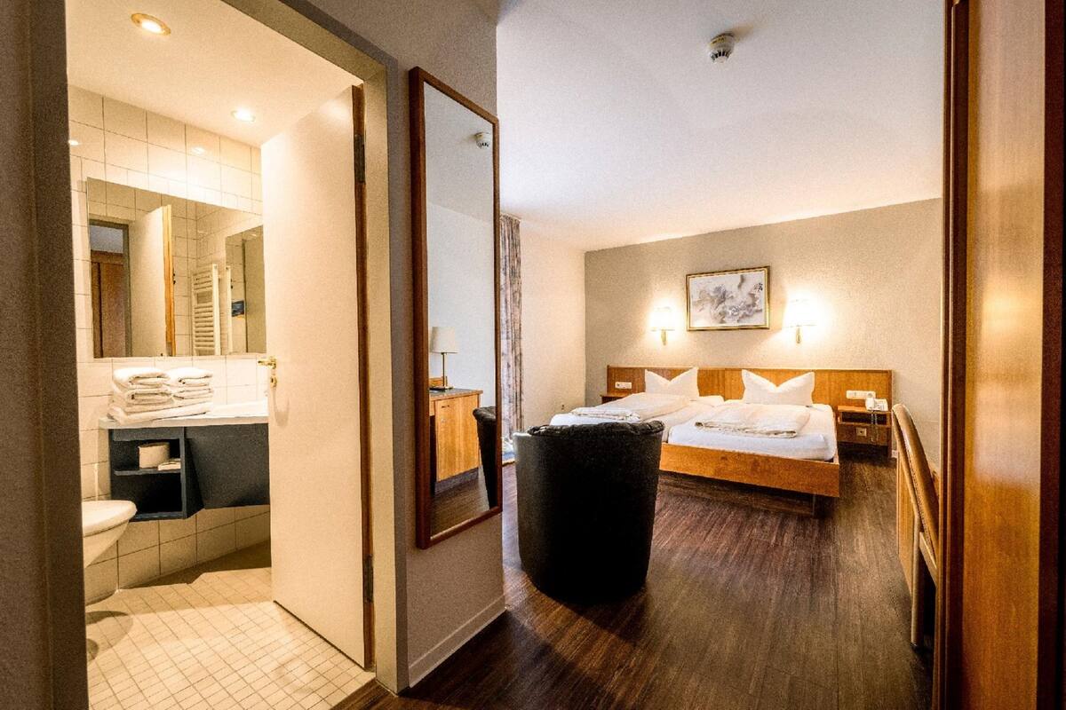 Ett hotellrum med trägolv, en säng med vita sängkläder, ett nattduksbord, en spegel och en väggmonterad lampa.