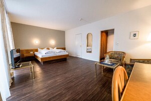 Habitación de hotel con cama, escritorio, silla y una mesita. Hay un espejo en la pared y una puerta que da a otra habitación.