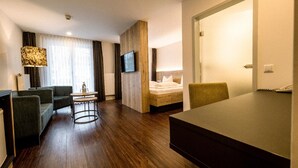 A hotel room with a wooden floor, a sofa, a small table, a bed, and a TV mounted on the wall. - Kurhotel San Andreas (Bad Fuessing)