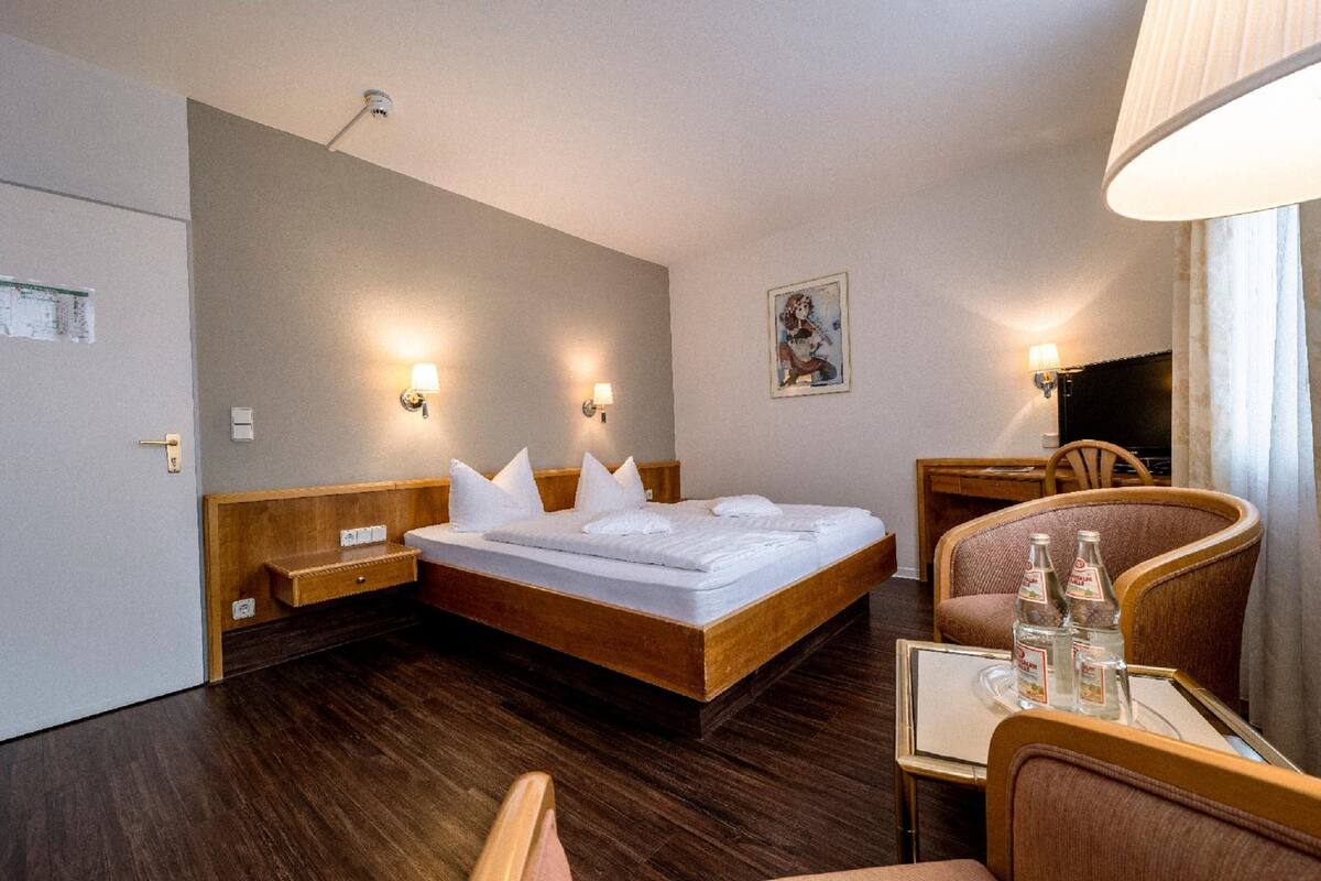 Ett hotellrum med en säng i trä, sängbord, ett litet bord med flaskor och en stol.
