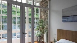 Ein Schlafzimmer mit Bett, Nachttisch, einem Topfpflanzen und Blick durch eine Glastür auf eine begrünte Außenanlage.