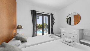 A hotel room with two beds, a large window with curtains, a round mirror, a bedside table, and a view of a swimming pool.