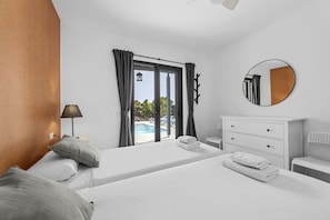 Ein Hotelzimmer mit zwei Betten, einem großen Fenster mit Vorhängen, einem runden Spiegel, einem Nachttisch und Blick auf einen Swimmingpool.