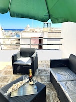 Una terraza en la azotea con mobiliario de mimbre, una botella de champán y vistas al mar y edificios.