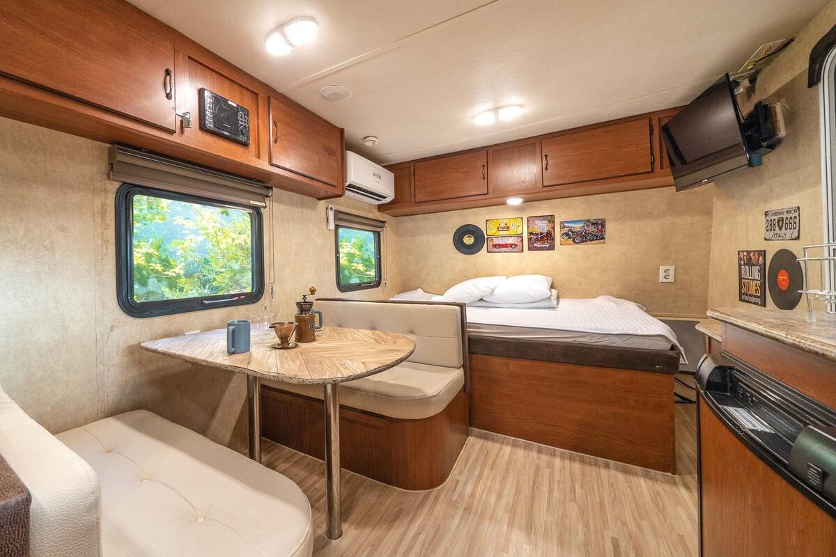 A compact RV interior with a kitchenette, a dining area, a bed, and a window with a view of greenery.