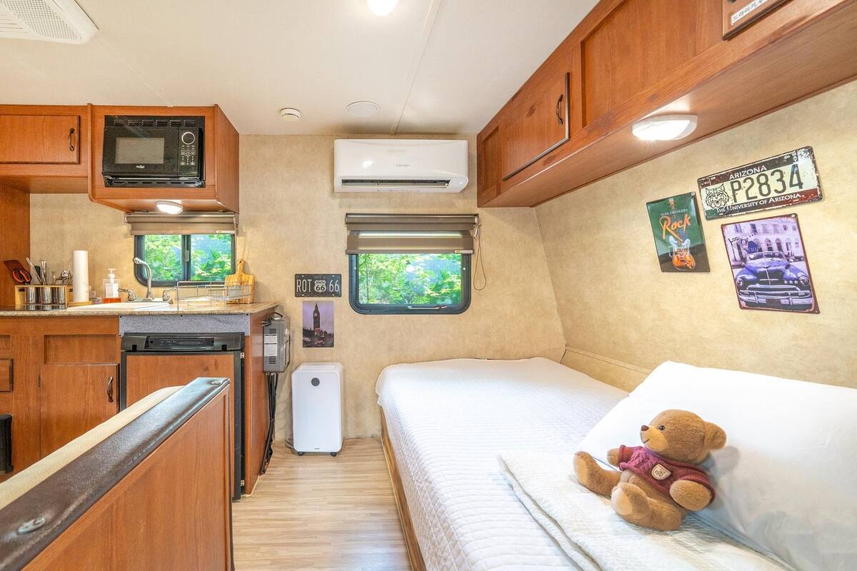 A compact RV kitchen with a microwave, sink, and compact refrigerator. There's a bed with a teddy bear and a window with a view of greenery.