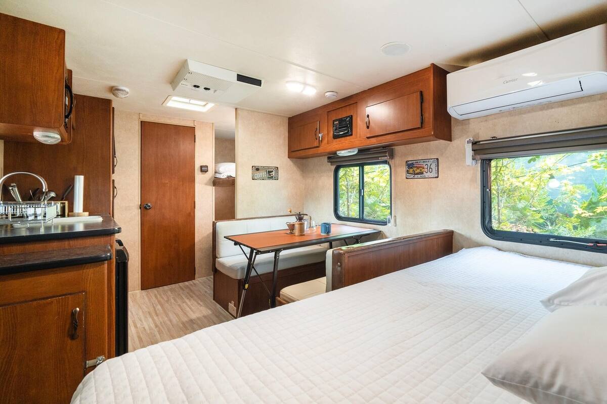 A compact RV interior with a kitchenette, a dining area, a bed, and a window with a view of greenery.