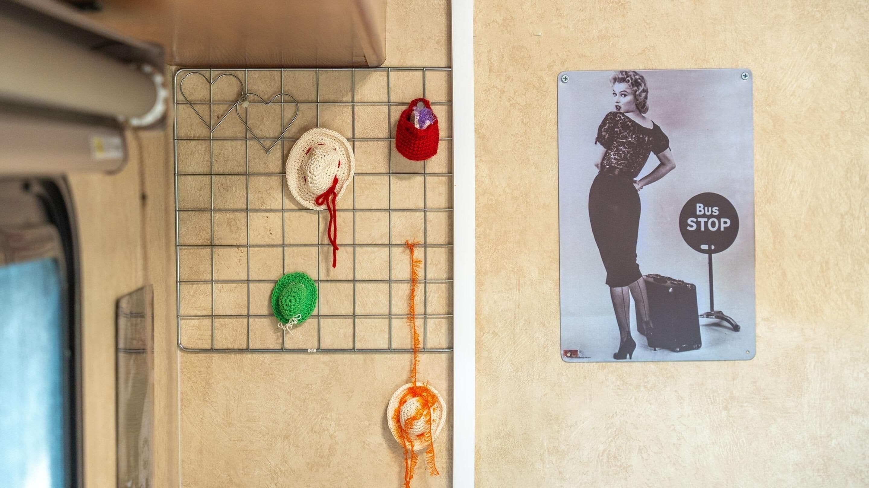 A kitchen area with a metal rack holding various items, a wall-mounted sign reading "Bus Stop," and a vintage advertisement featuring a woman with luggage.
