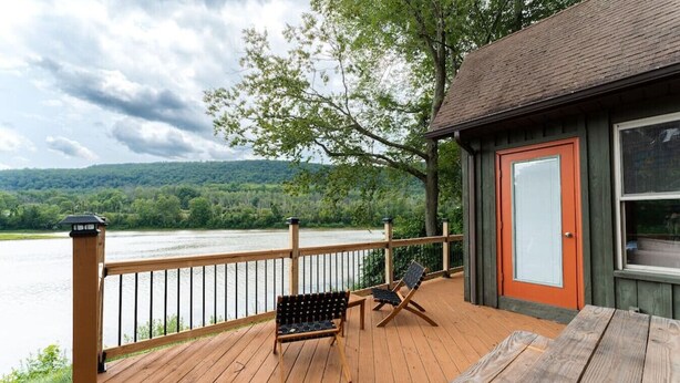 Una terraza de madera con vista a un río y montañas, un pequeño edificio con una puerta naranja y dos sillas.