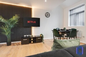 Una sala moderna con un televisor de pantalla plana mostrando "NETFLIX", un comedor con mesa y sillas, y un ventanal con persianas.