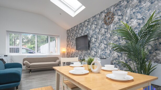 A modern living room with a wooden dining table set for four, a sofa, a TV mounted on the wall, and a large window with a view of a car outside. - Santorini Blues by CEFAS - Free Parking - Bungalow (Greater Manchester)