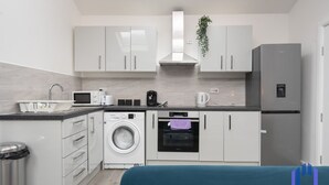 A compact kitchen with white cabinets, a washing machine, an oven, and a refrigerator. - Santorini Blues by CEFAS - Free Parking - Bungalow (Greater Manchester)