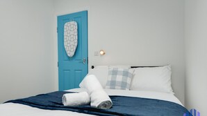 A hotel room with a blue door, a bed with white bedding, a blue blanket, and a checkered pillow. - Santorini Blues by CEFAS - Free Parking - Bungalow (Greater Manchester)
