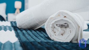 A neatly folded white towel with a label on a bed with a blue textured cover. - Santorini Blues by CEFAS - Free Parking - Bungalow (Greater Manchester)