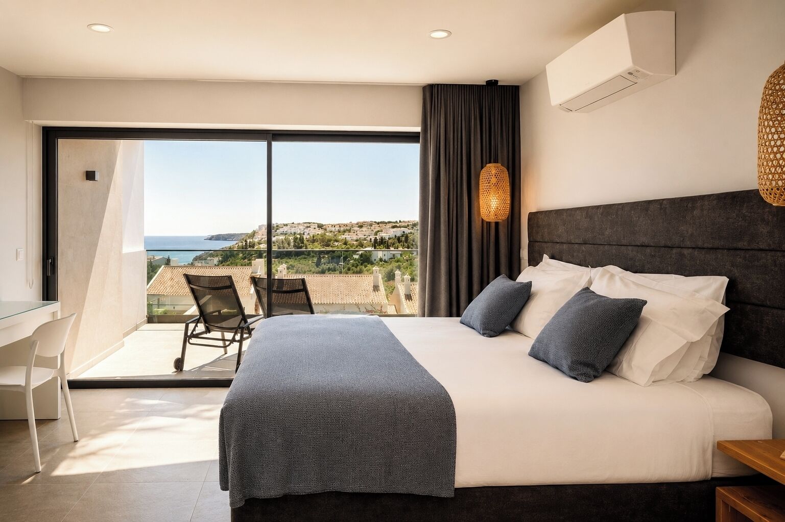 Une chambre avec un grand lit, un bureau et une chaise, un balcon avec vue sur la mer et un luminaire suspendu.