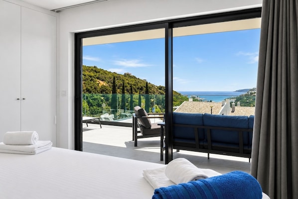 Quarto de hotel moderno com uma grande porta de vidro deslizante que dá para uma varanda com vista para a paisagem costeira.