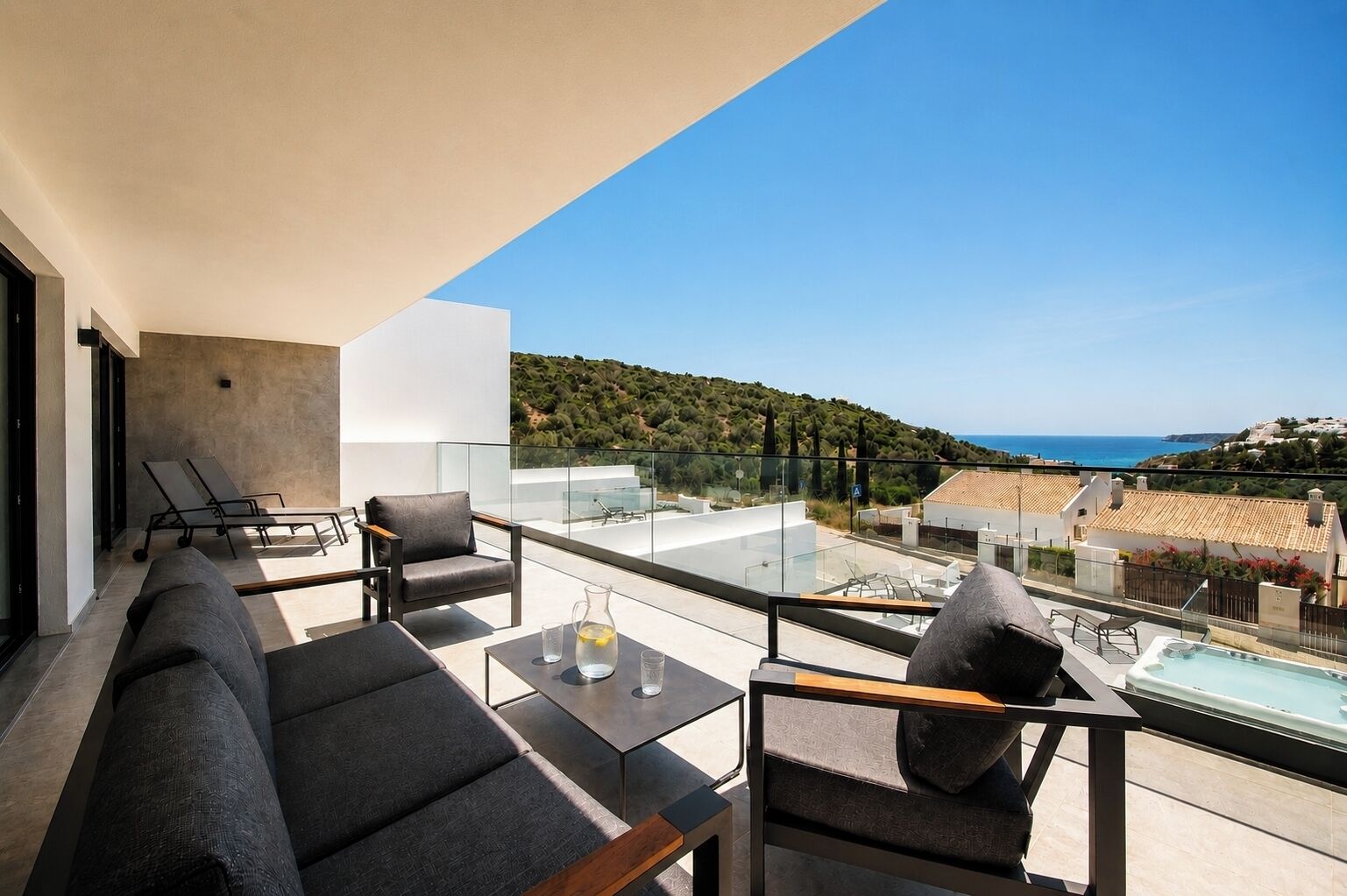 Ein Balkon mit einer Couch, Stühlen, einem Tisch und Blick auf Meer und Hügel.
