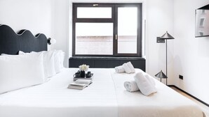 Una habitación de hotel con una cama, una ventana con vistas, una mesita de noche con un libro y un florero, y una lámpara.