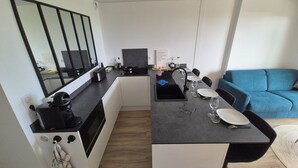 Een compacte keuken met een modern design, voorzien van een ingebouwde oven, een magnetron en een spoelbak. Er is een eethoek met een tafel voor twee personen, en een blauwe bank in de woonkamer.