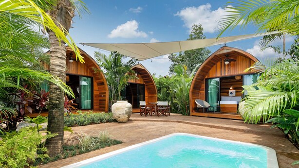 Uma área de piscina com duas cabanas de madeira em arco, rodeada por vegetação exuberante e um céu azul límpido.
