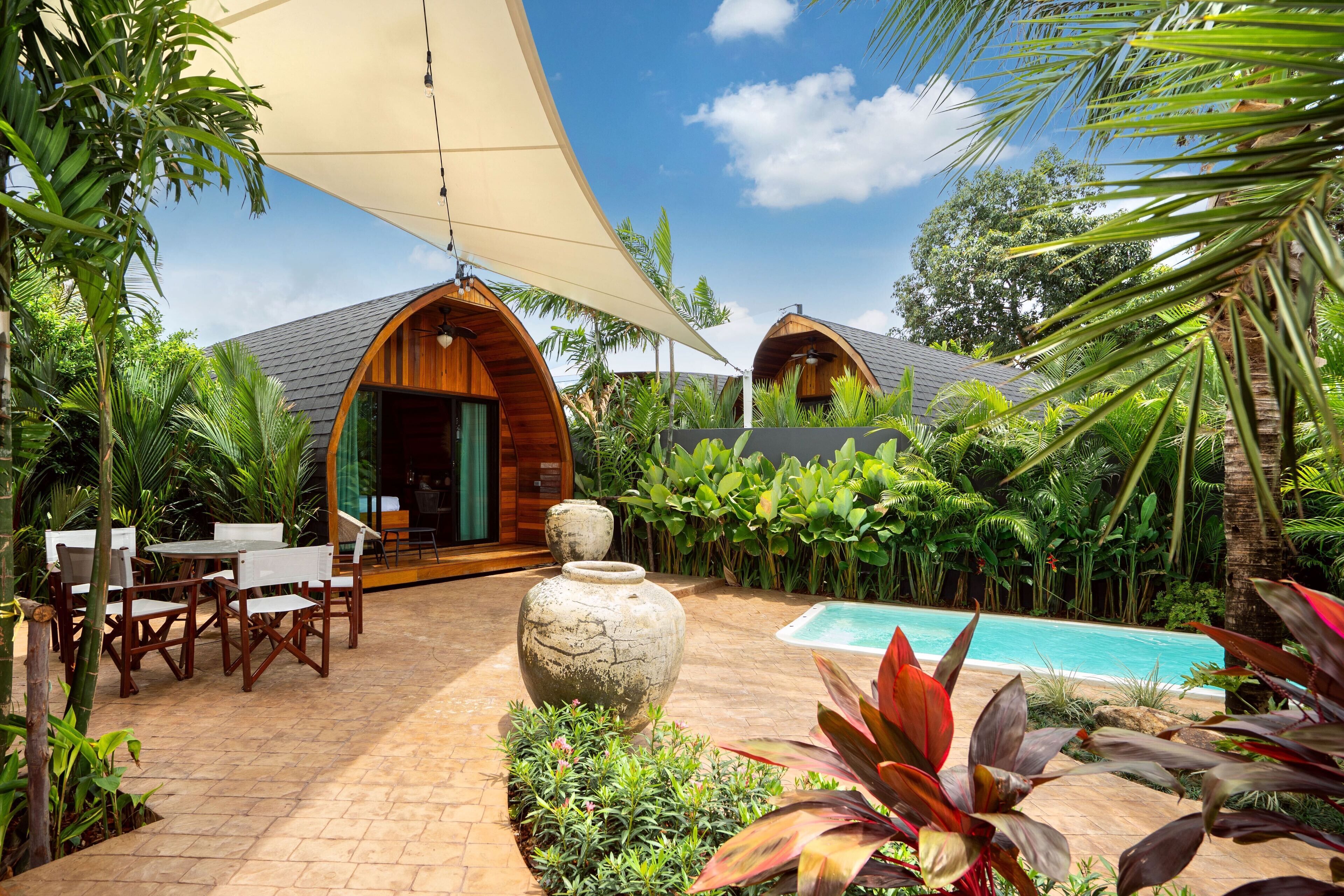 Un espace aménagé au bord de la piscine, avec des cabanons en bois, un ensemble de mobilier de jardin et une végétation tropicale luxuriante.