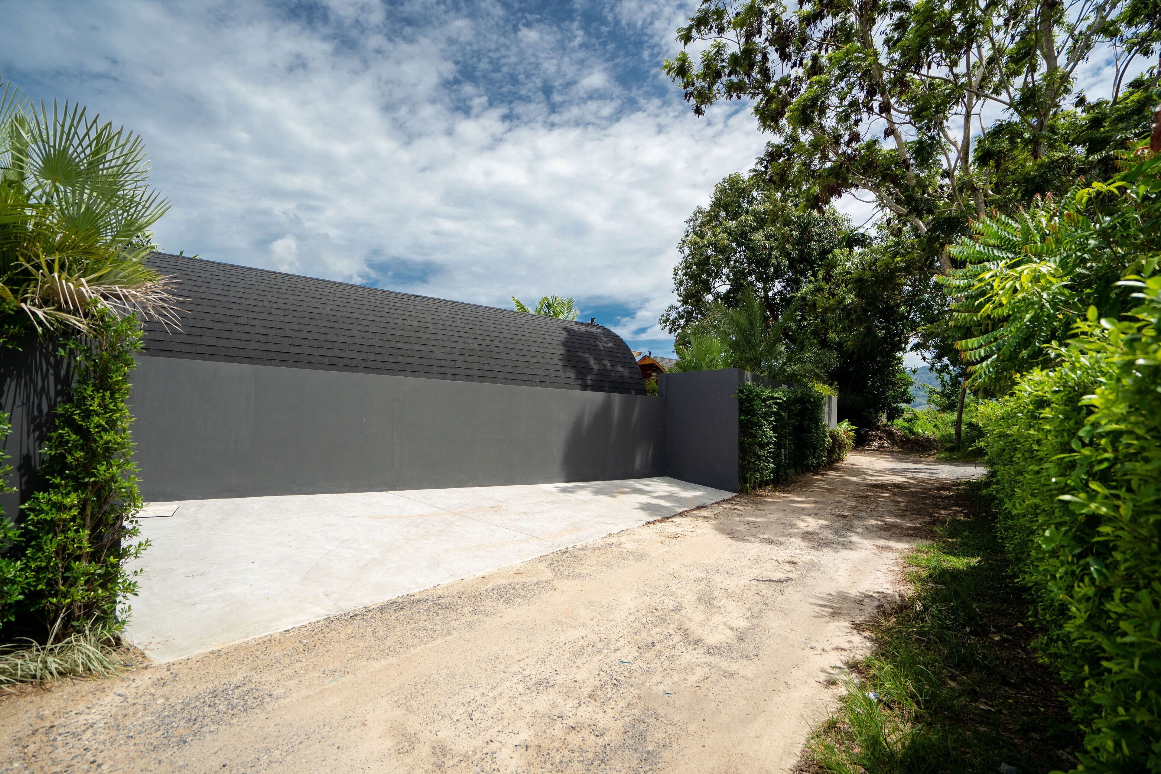 A modern building with a grey facade, a concrete driveway, and lush greenery surrounding it.