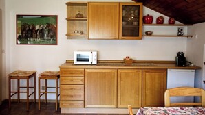 Eine kleine Küche mit Holzschränken, einer Mikrowelle und einem kleinen Kühlschrank. Zwei Hocker und ein Stuhl sind vorhanden.