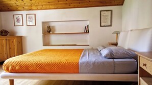 Ein Zimmer mit einem Einzelbett, Holzboden, einem weißen Bett mit orangefarbener Decke, einem Nachttisch aus Holz mit Lampe sowie einem Regal mit Büchern und einer Vase.