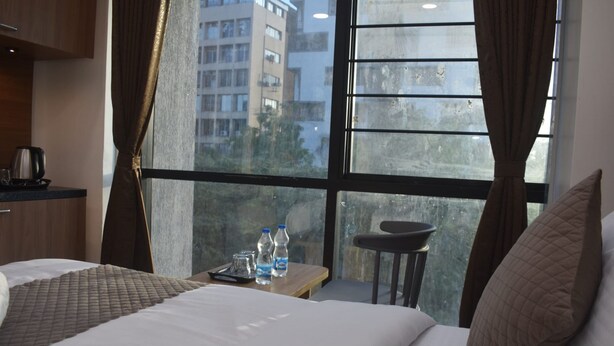 A hotel room with a large window, a bed with a quilted cover, a small table with water bottles, and a chair. - Hotel Green Fortune (Ahmedabad)