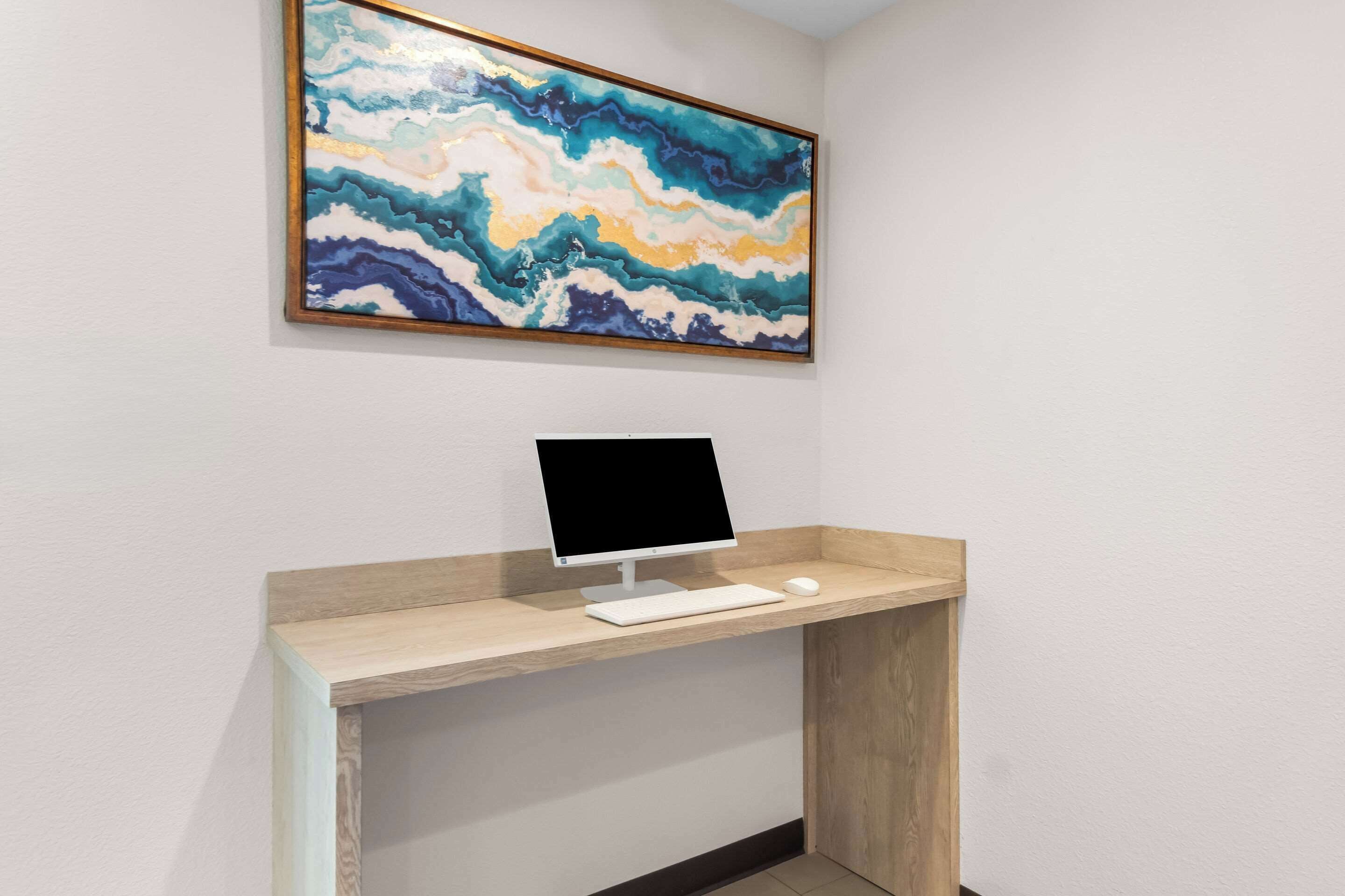 Un bureau d’ordinateur avec un écran, un clavier et une souris, et une peinture abstraite aux couleurs vives accrochée au-dessus.