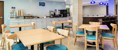 Un café moderne avec des tables en bois et des chaises rembourrées bleues, un comptoir où sont exposés les plats, et un motif hexagonal bleu sur un mur.