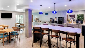 A modern hotel kitchen with bar stools, a long counter, and blue pendant lights. - Clarion Pointe Fort Worth East - Arlington (Fort Worth)