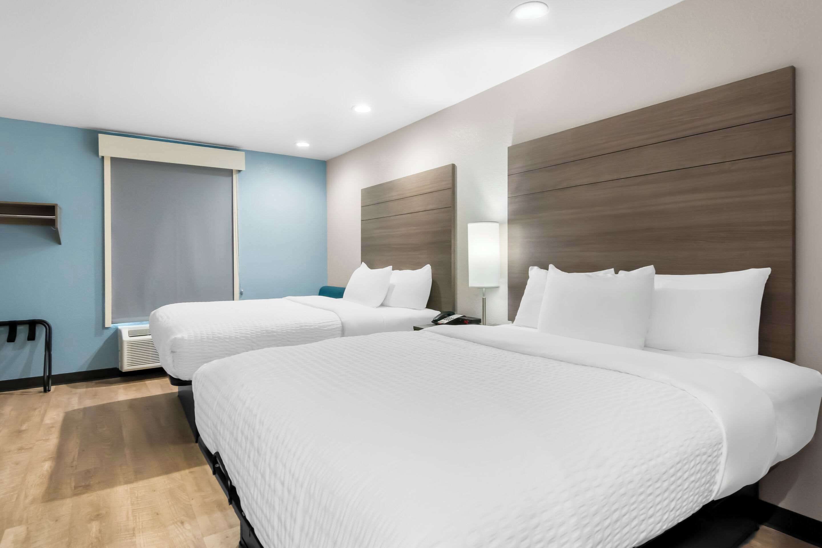 Habitación de hotel con dos camas, un ventanal grande, una mesita de noche con lámpara y un televisor montado en la pared.