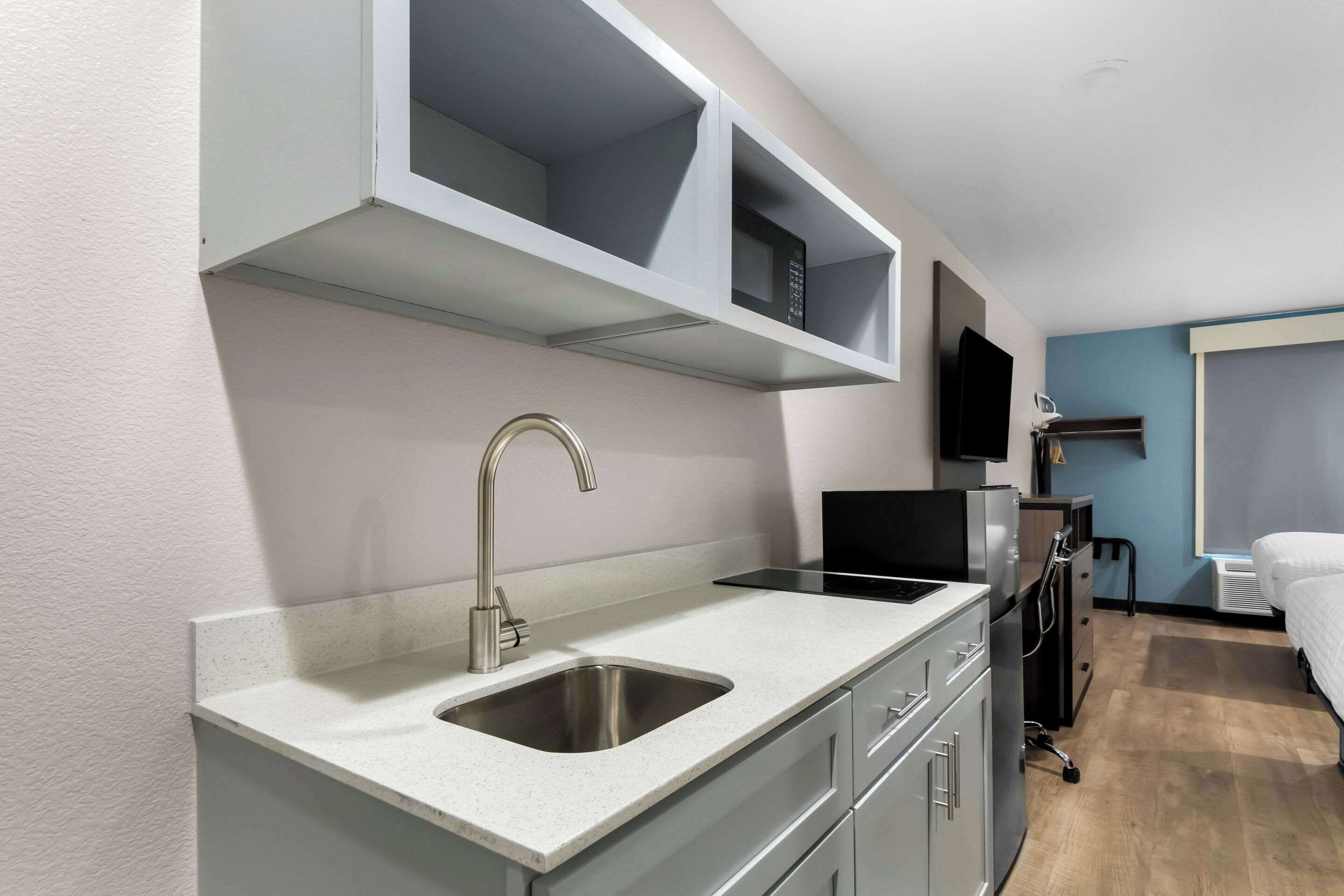 A modern kitchenette with a stainless steel sink, a built-in oven, and a microwave. There is a flat-screen TV mounted on the wall above the oven.