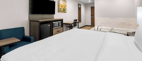 A hotel room with a large bed, a desk with a flat-screen TV, a microwave, a chair, a small table, and a bathroom with a bathtub and sink.