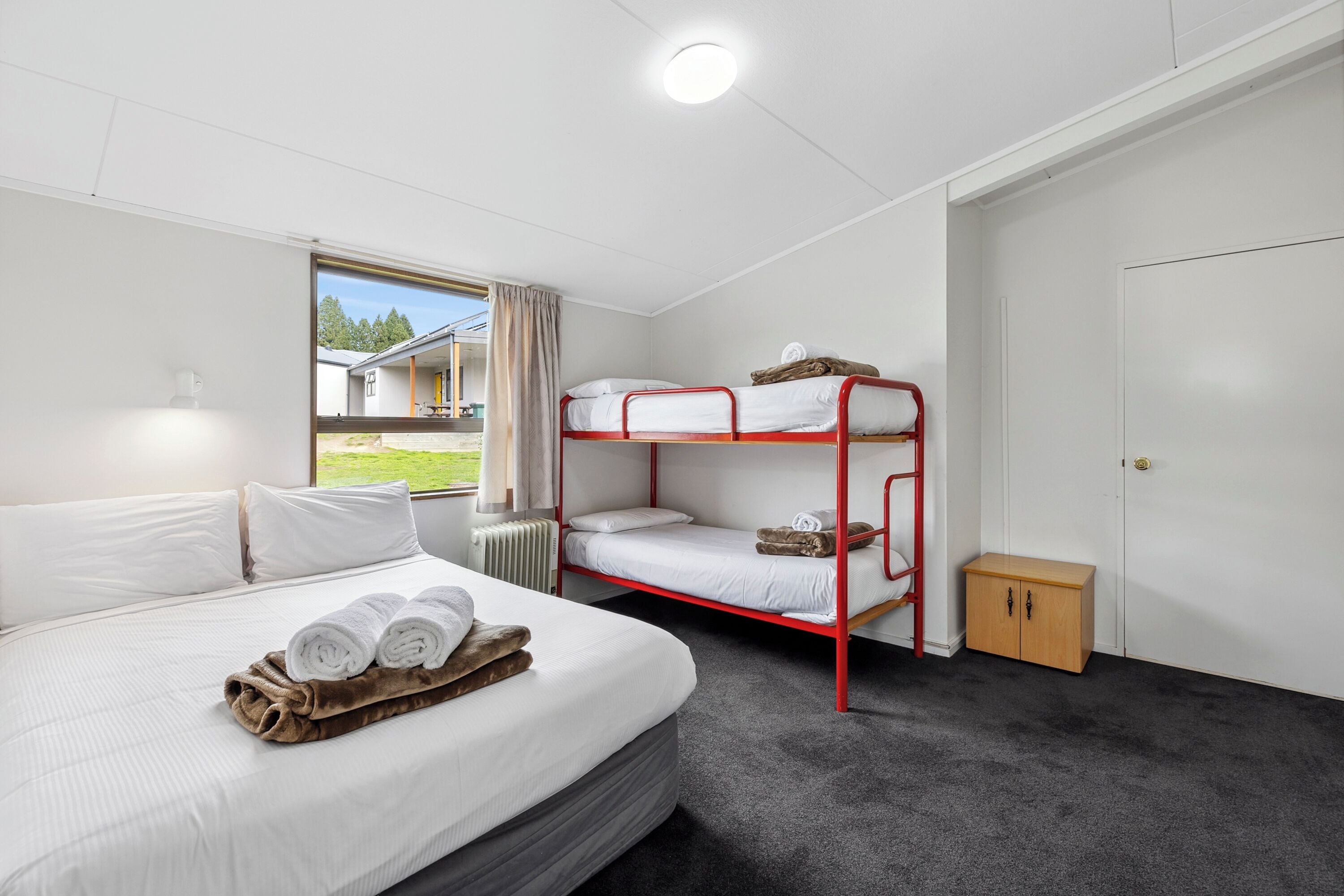 A room with a single bed, a bunk bed, a small wooden cabinet, and a window with a view of a building and greenery.