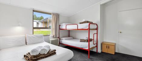 A room with a single bed, a bunk bed, a small wooden cabinet, and a window with a view of a building and greenery.