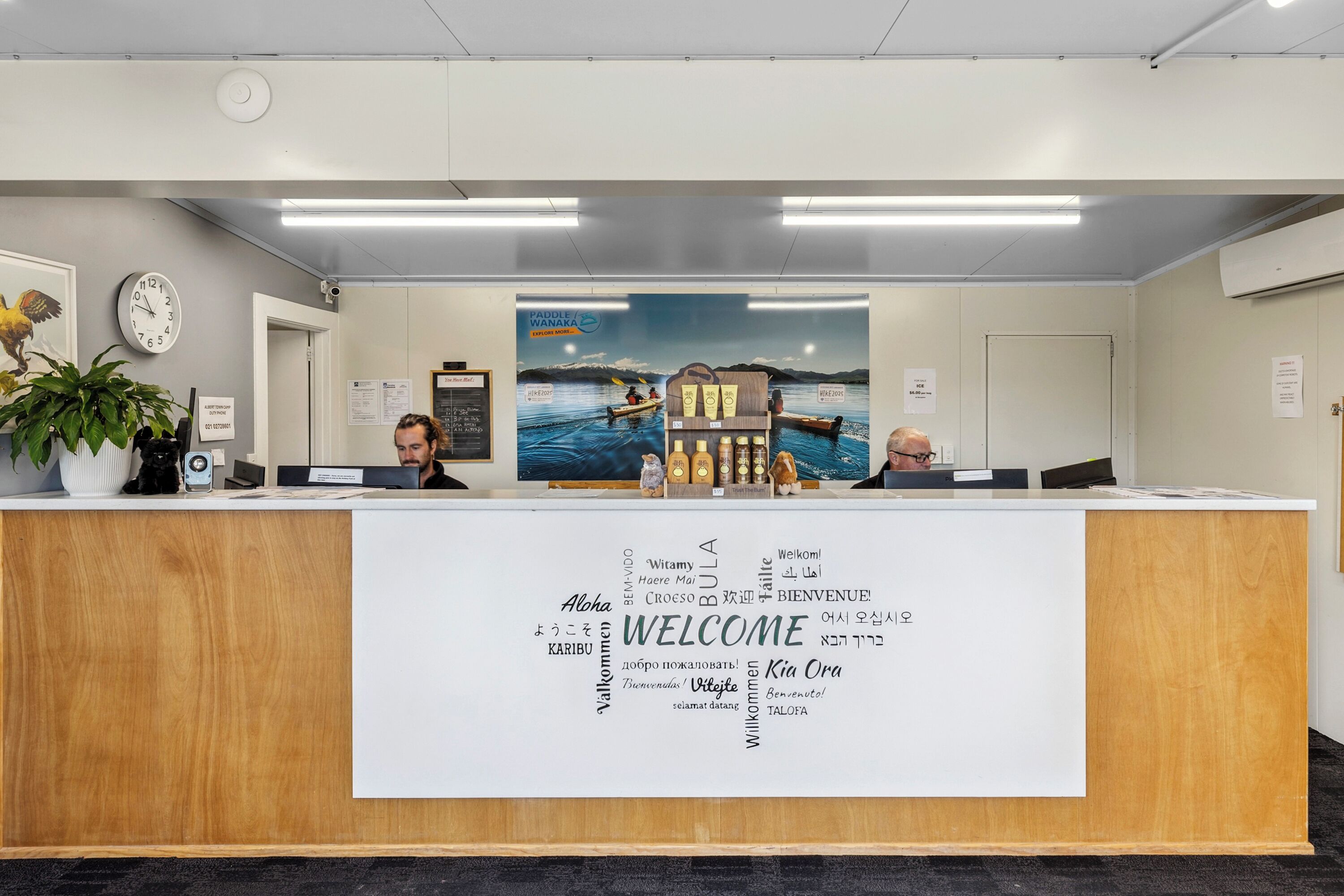 A reception desk with a welcome sign featuring multiple languages, a clock, and a plant.