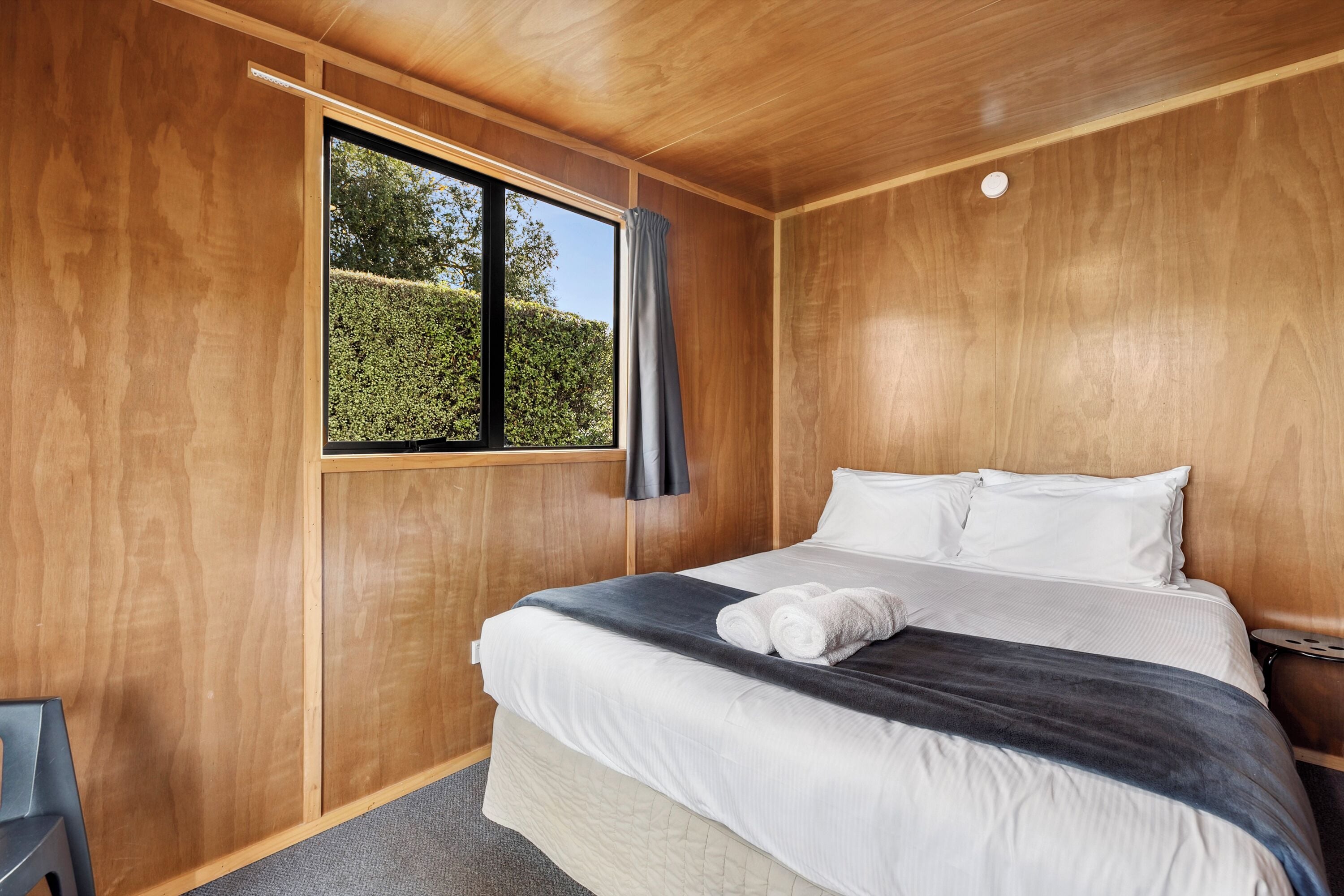 Un dormitorio con una cama grande, paredes de madera, una ventana con vistas a zonas verdes y una silla gris.