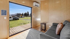 Un interno moderno con un divano grigio, un tavolino rotondo e un'ampia porta scorrevole in vetro che si apre su uno spazio esterno con un veicolo e una casa.