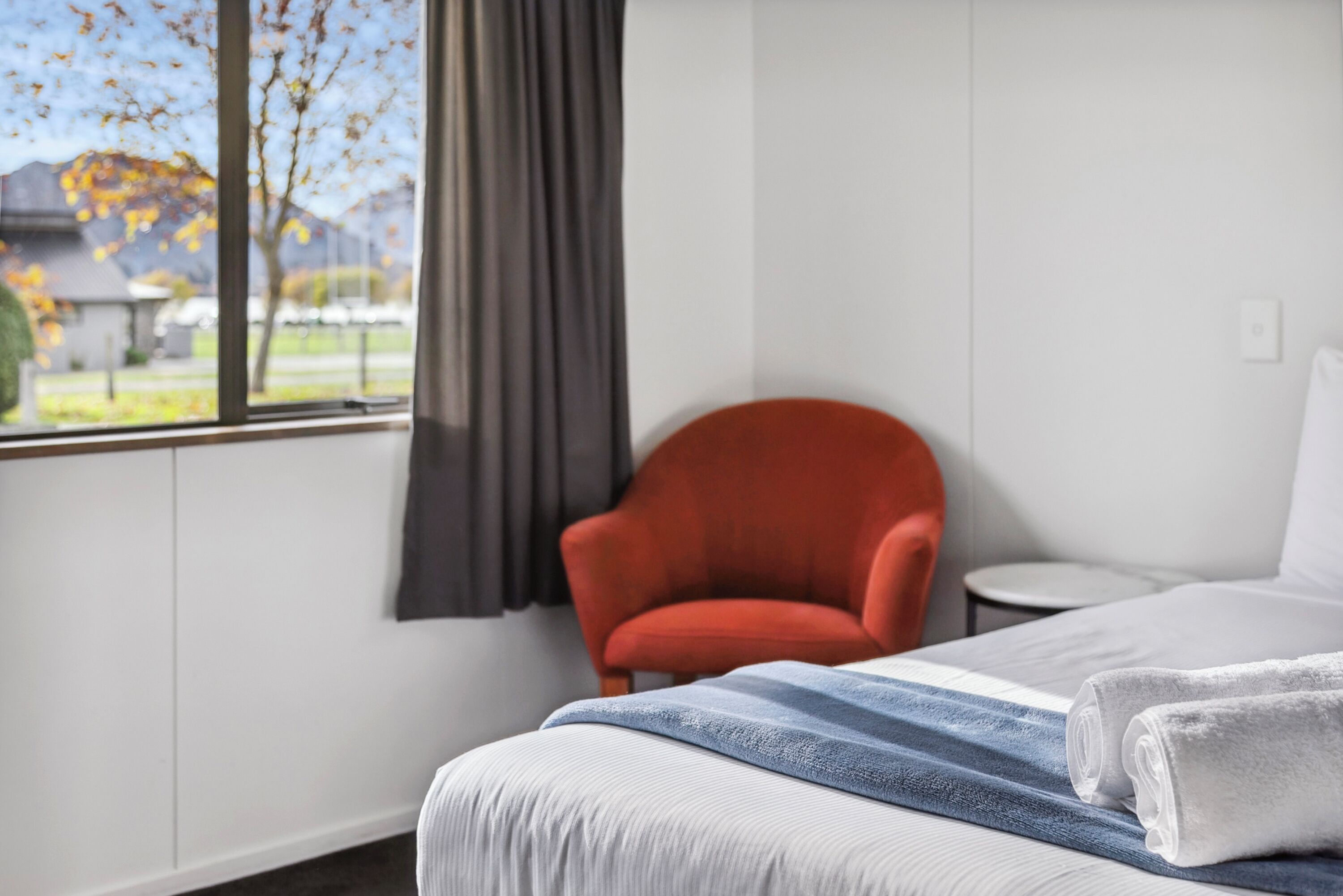 Habitación de hotel con cama, un sillón rojo, una mesita redonda y vistas a árboles y edificios a través de la ventana.