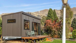 Una moderna e compatta cabina con veranda e posti a sedere all'aperto, immersa in un giardino curato con le montagne sullo sfondo.