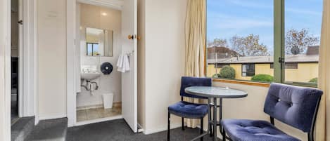 Habitación de hotel con baño, una mesa redonda, dos sillas y un ventanal con vistas al exterior.
