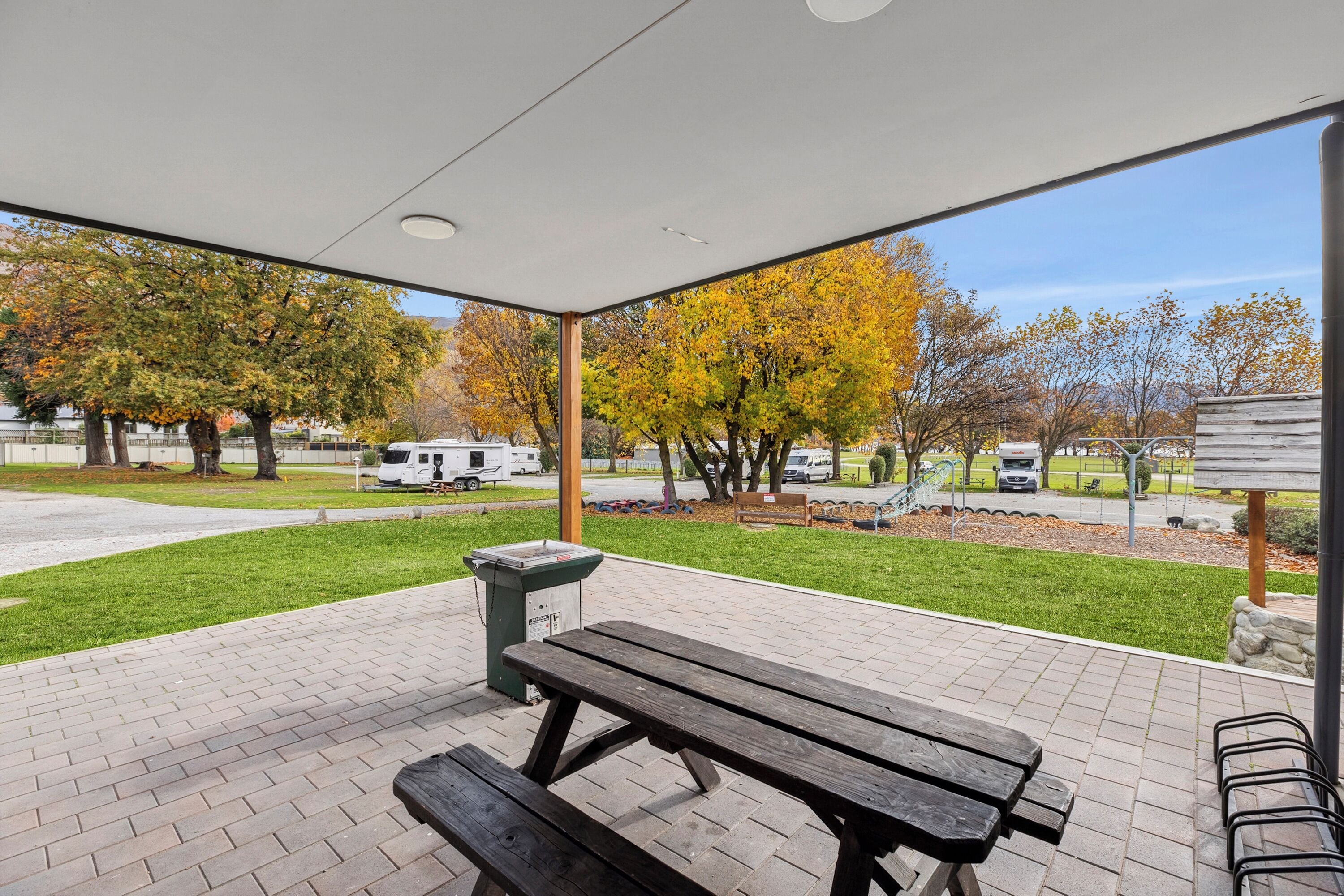 Un patio al aire libre con mesas de picnic y bancos, un bote de basura y una vista de árboles y vehículos al fondo.