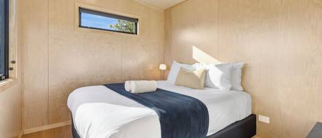 A neatly made bed with white and navy bedding, a small window with a view of trees, and a wooden floor.