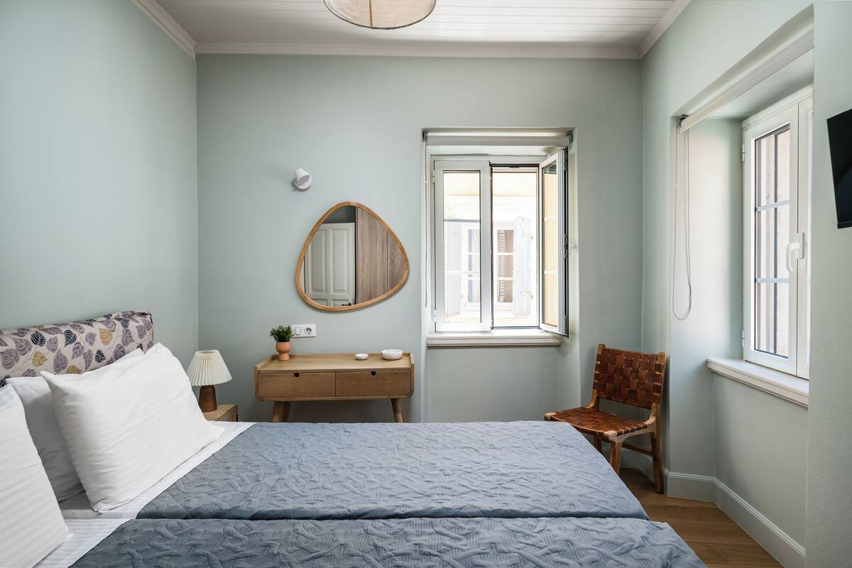 A bedroom with a bed, a wooden nightstand, a mirror, a window, and a chair.