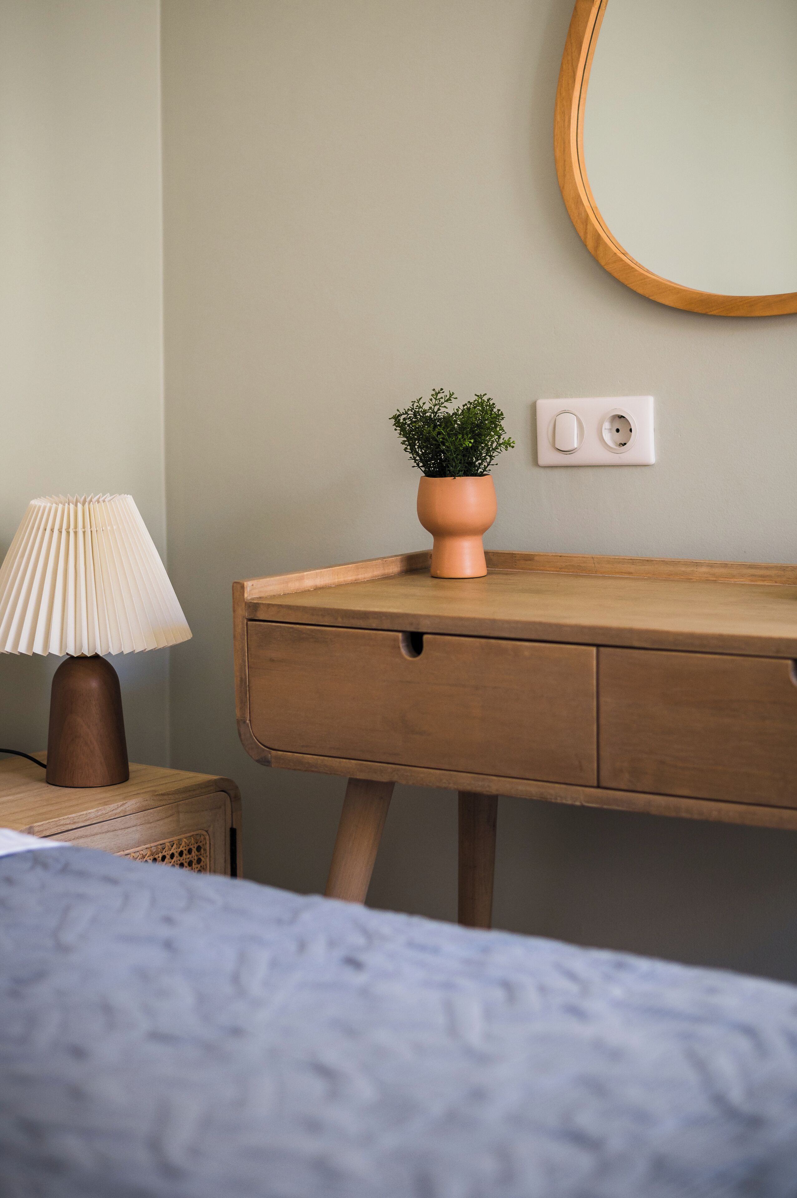A wooden bedside table with a lamp, a small potted plant, and a wall socket.