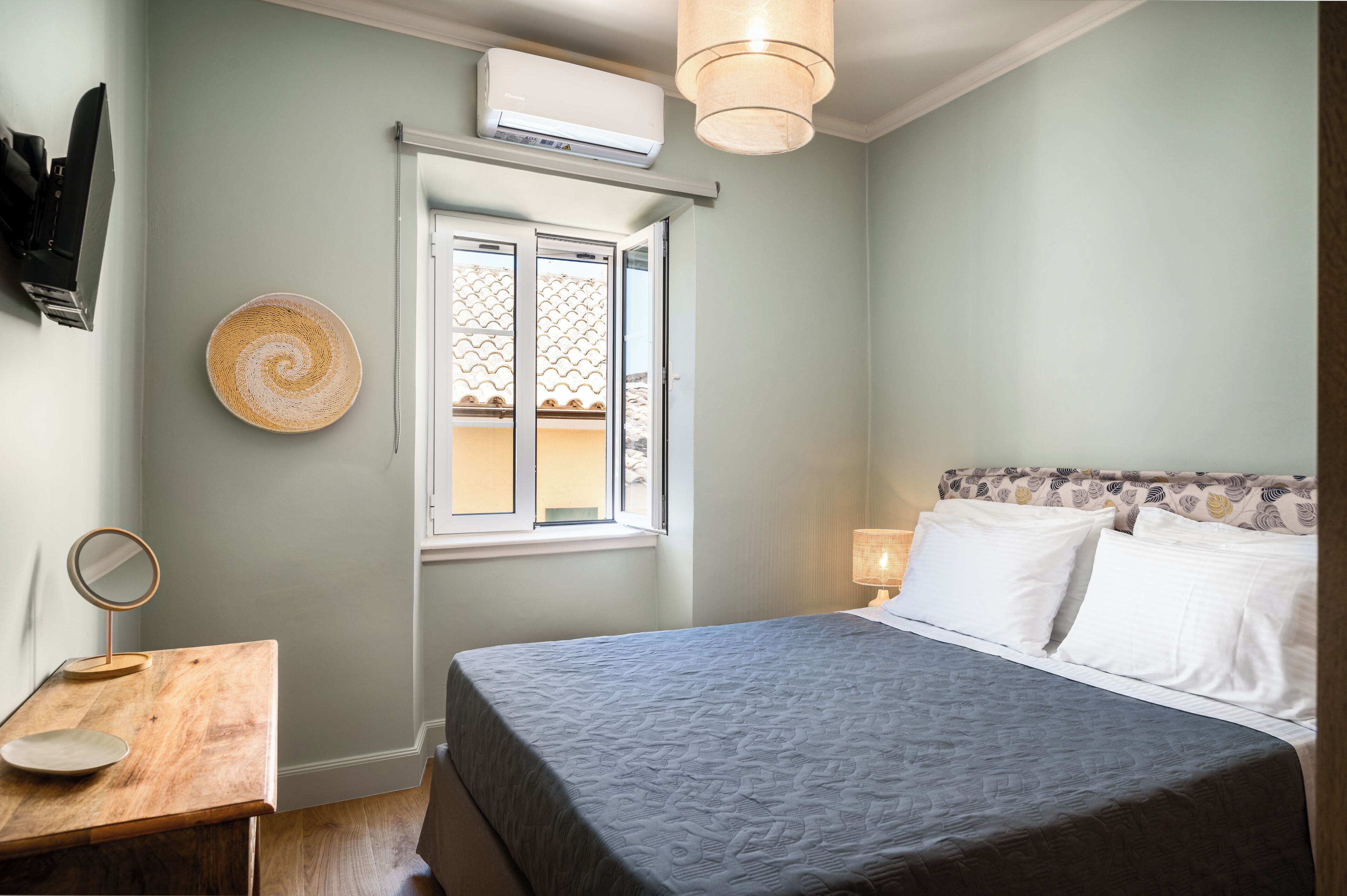 A bedroom with a bed, bedside table, wall-mounted air conditioner, window, and a decorative plate.