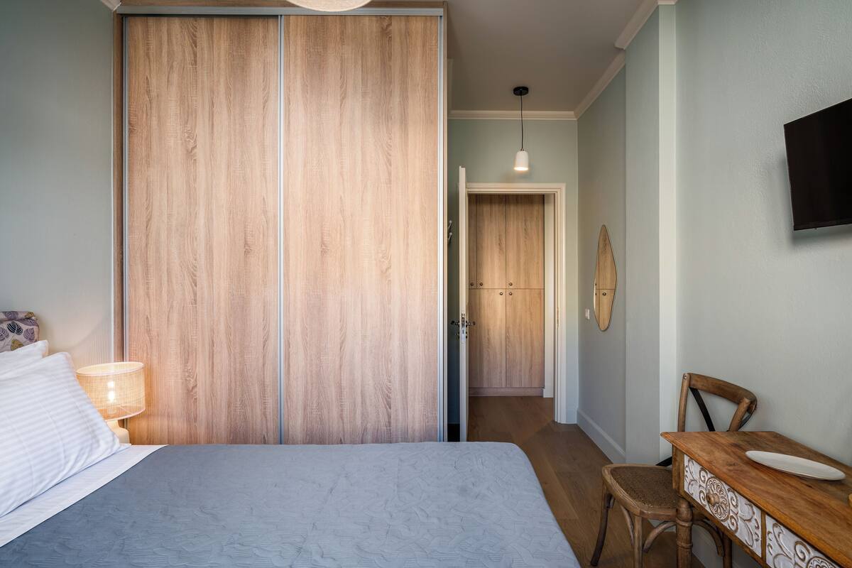 A bedroom with a bed, a wooden wardrobe, a desk with a chair, and a television on the wall.